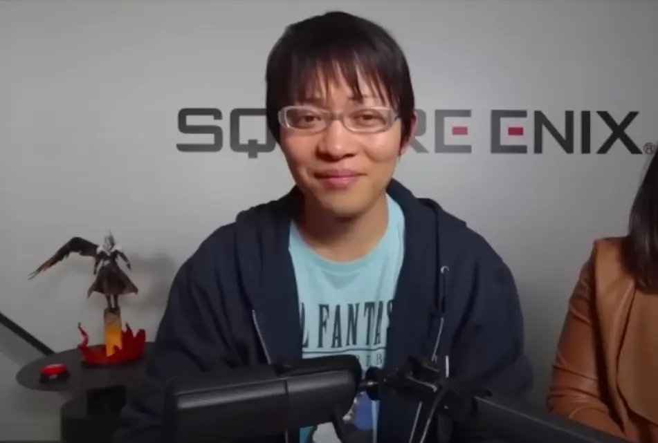 Naoki Hamaguchi iført briller og en blå t-shirt, siddende foran en Square Enix-væg.