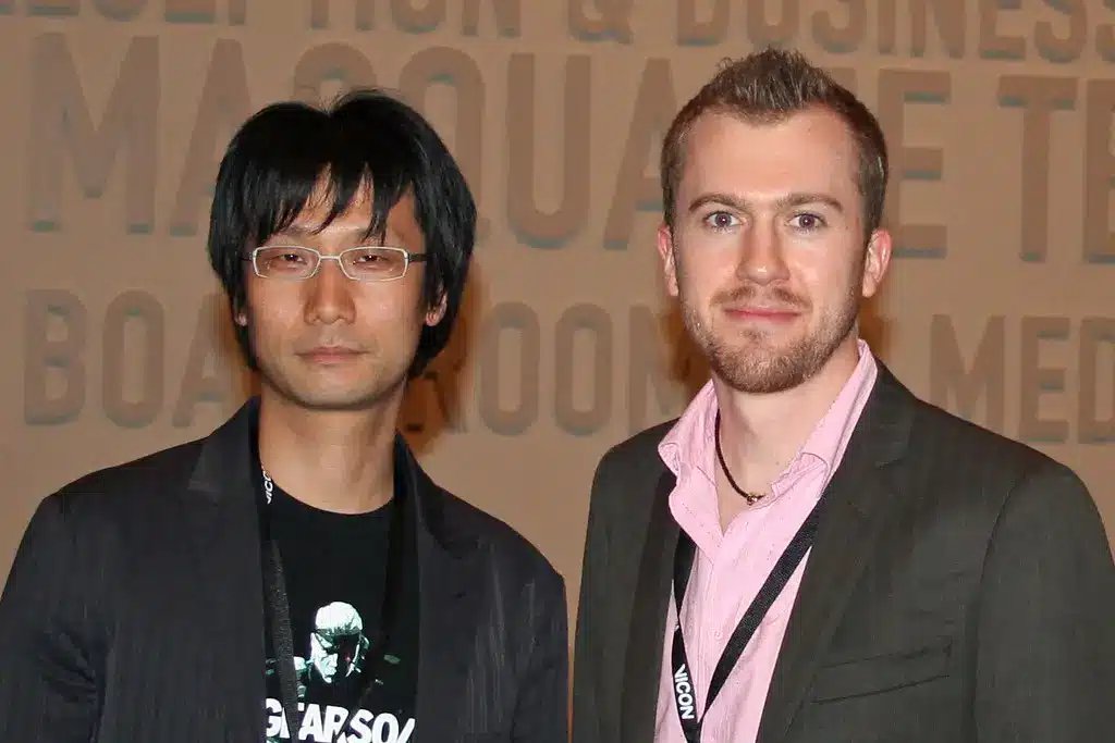 Hideo Kojima iført briller og en sort blazer, stående ved siden af en anden mand.
