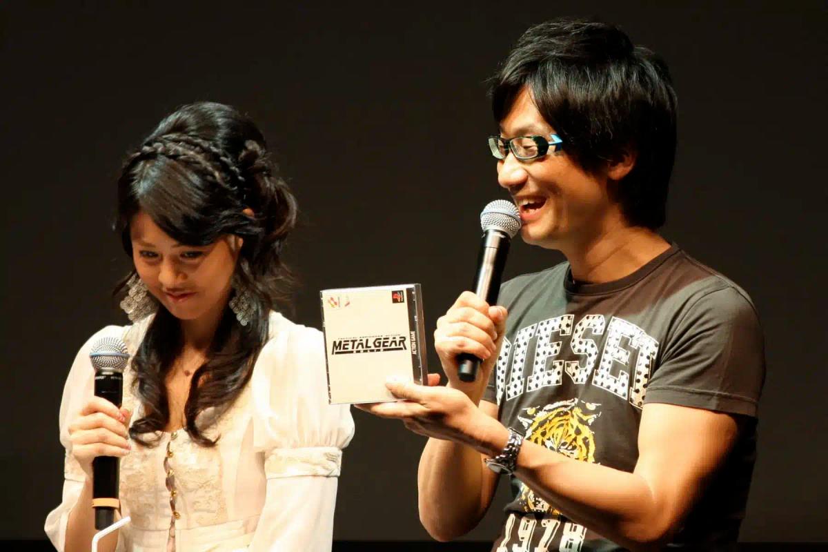 Hideo Kojima holder et Metal Gear-spilcover, mens han taler i en mikrofon.